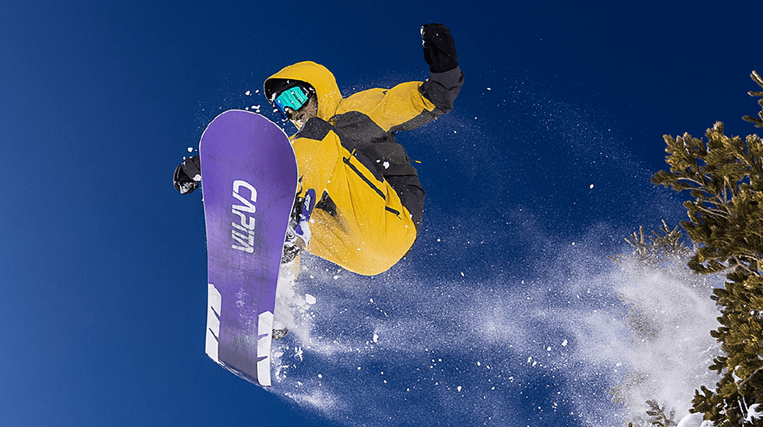 A snowboarder in a yellow jacket and purple board jumps high against a clear blue sky, with snow spraying.