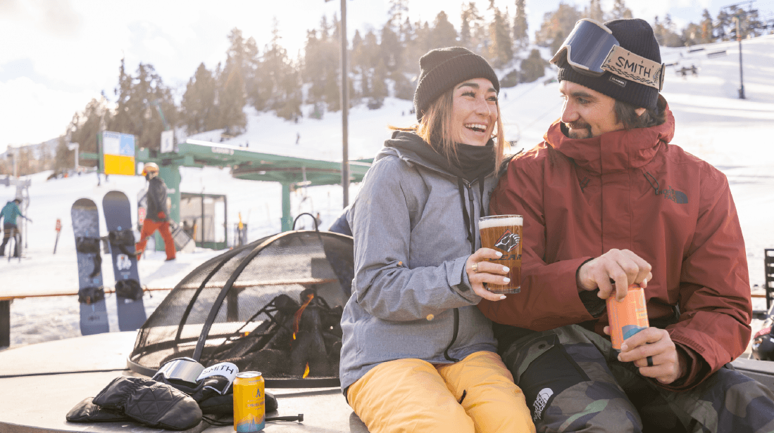 A laughing woman and a man in winter gear enjoying drinks by a fire pit at a sunny ski resort.