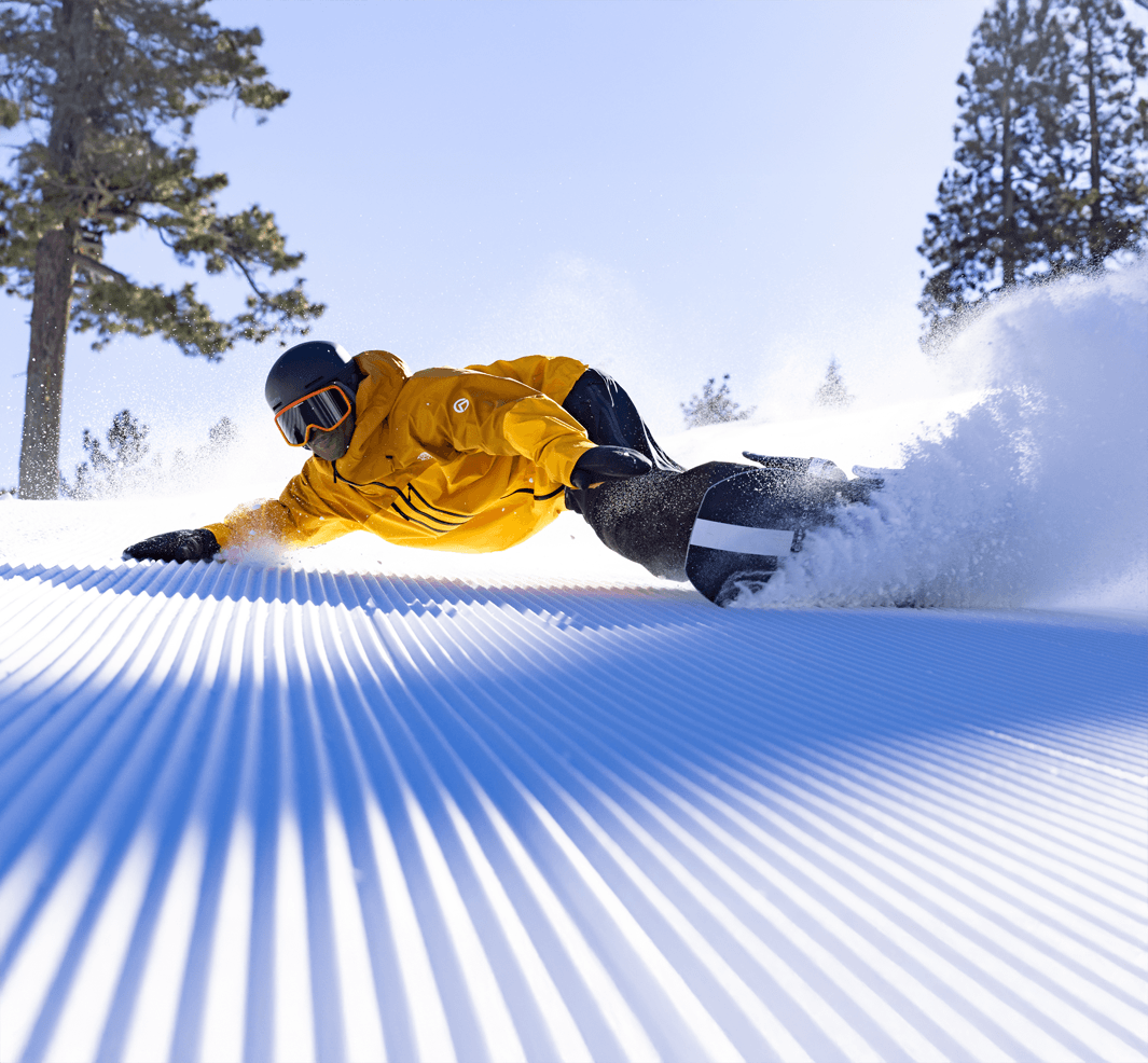 A snowboarder in a bright yellow jacket carves sharply on a groomed slope, kicking up a spray of snow.