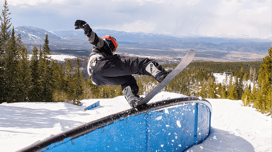 A snowboarder in dark gear and a red helmet performs a trick on a blue rail.